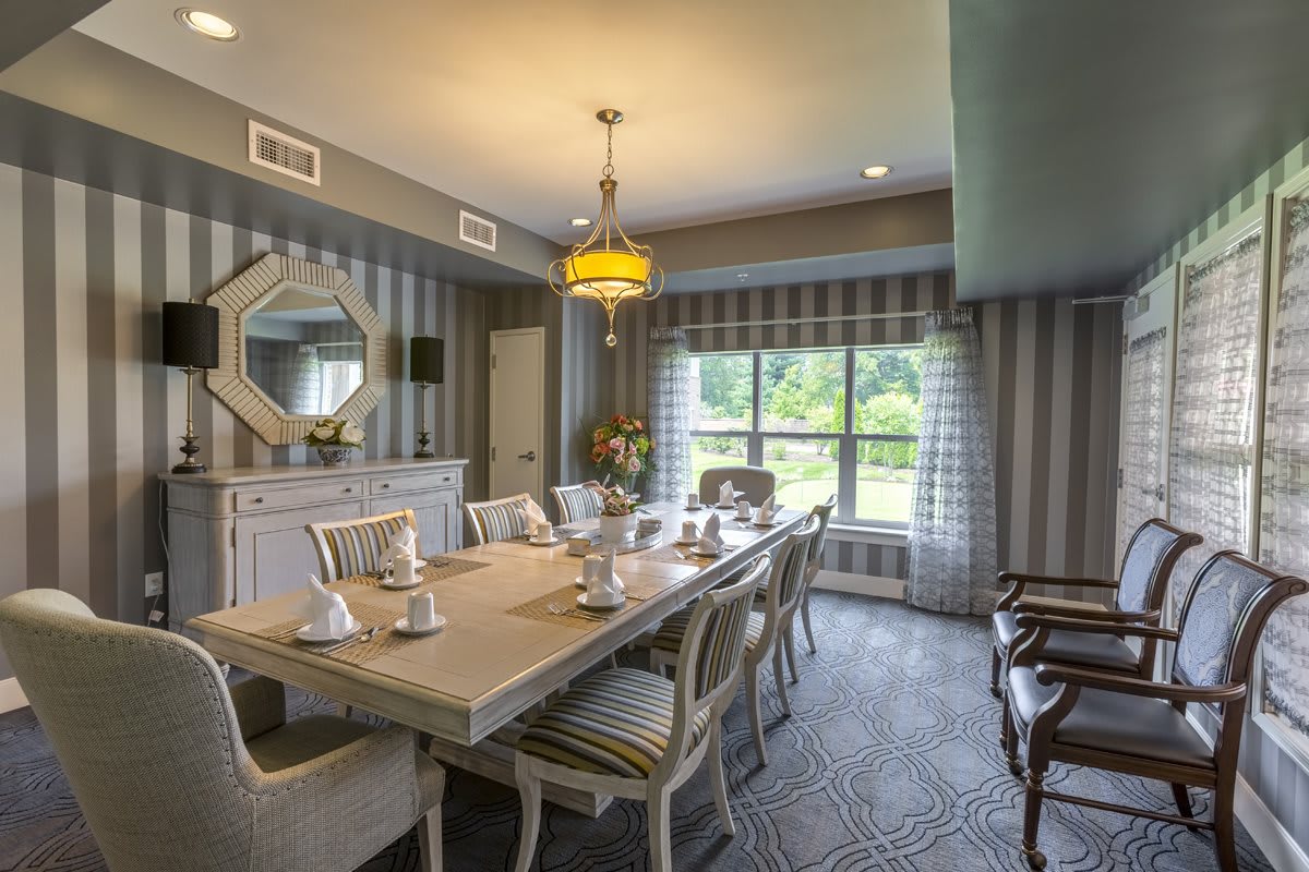 A stylish dining room with a long table and chairs, featuring striped wall decor and a large window providing natural light.