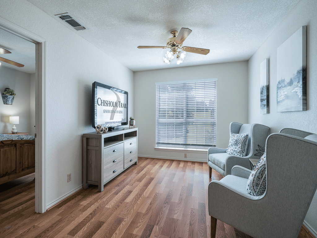 A modern living room with wooden flooring, featuring a television, chairs, and natural light from a large window.