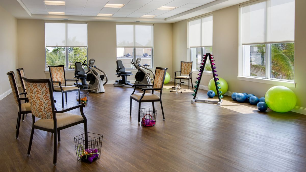 A well-lit exercise room equipped with stationary bikes, chairs, yoga balls, and a rack of dumbbells.