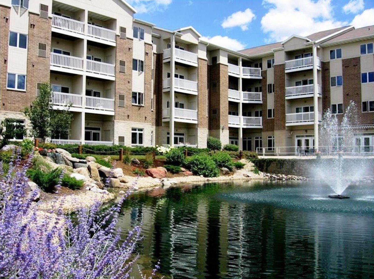 A modern apartment complex with balconies overlooks a serene pond and fountain.