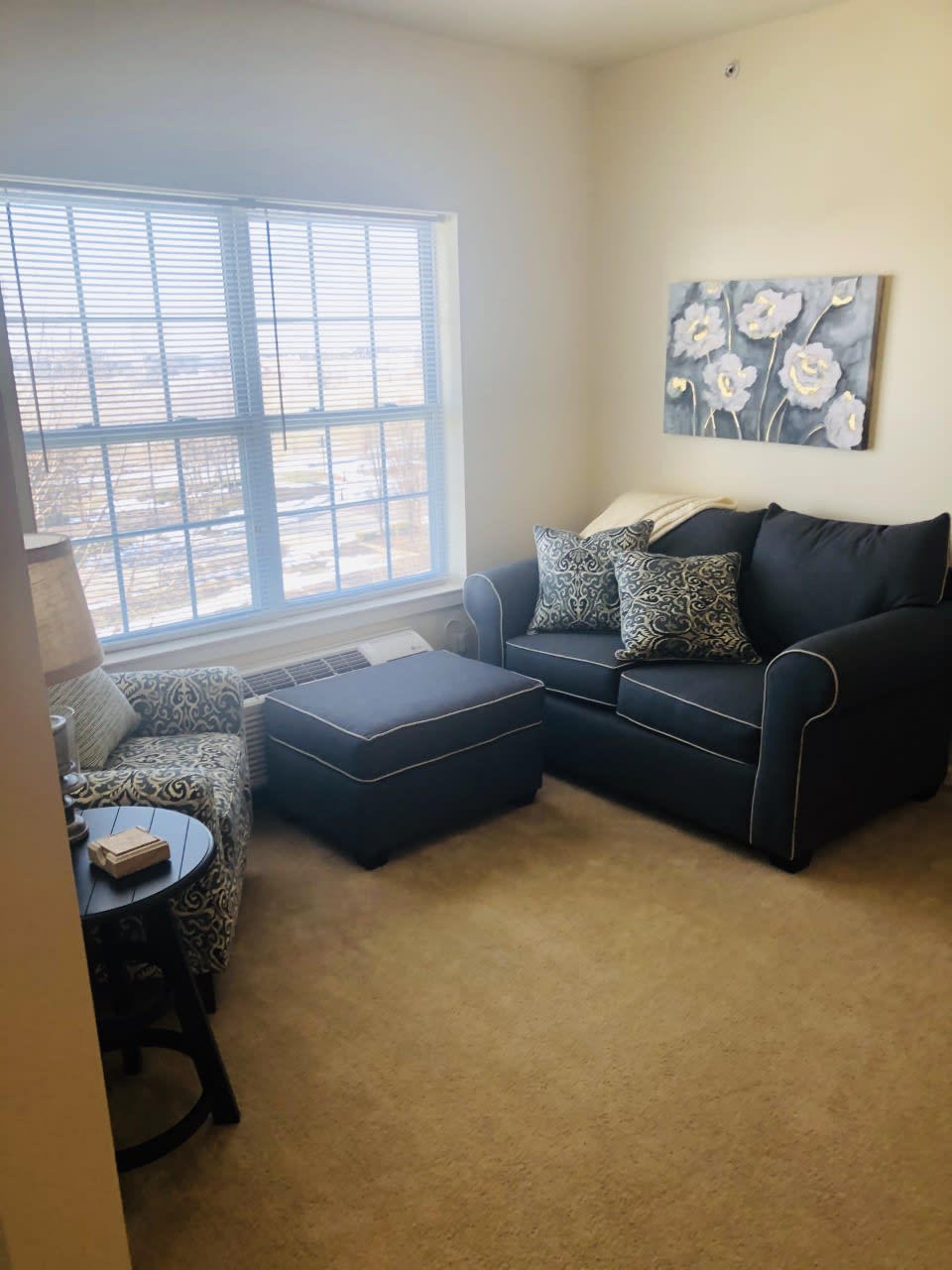 A cozy living room with a dark sofa, armchair, and ottoman, featuring a large window and floral artwork.