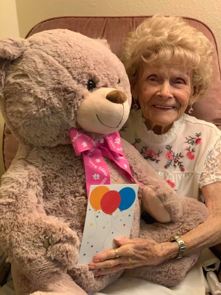 An elderly woman smiles while holding a large teddy bear and a gift card, suggesting a celebration or special occasion.