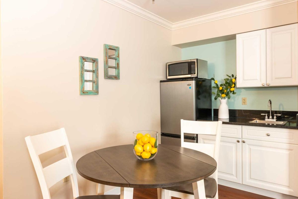 A compact kitchen area with a round table and chairs, featuring a stainless steel refrigerator and a bowl of lemons.