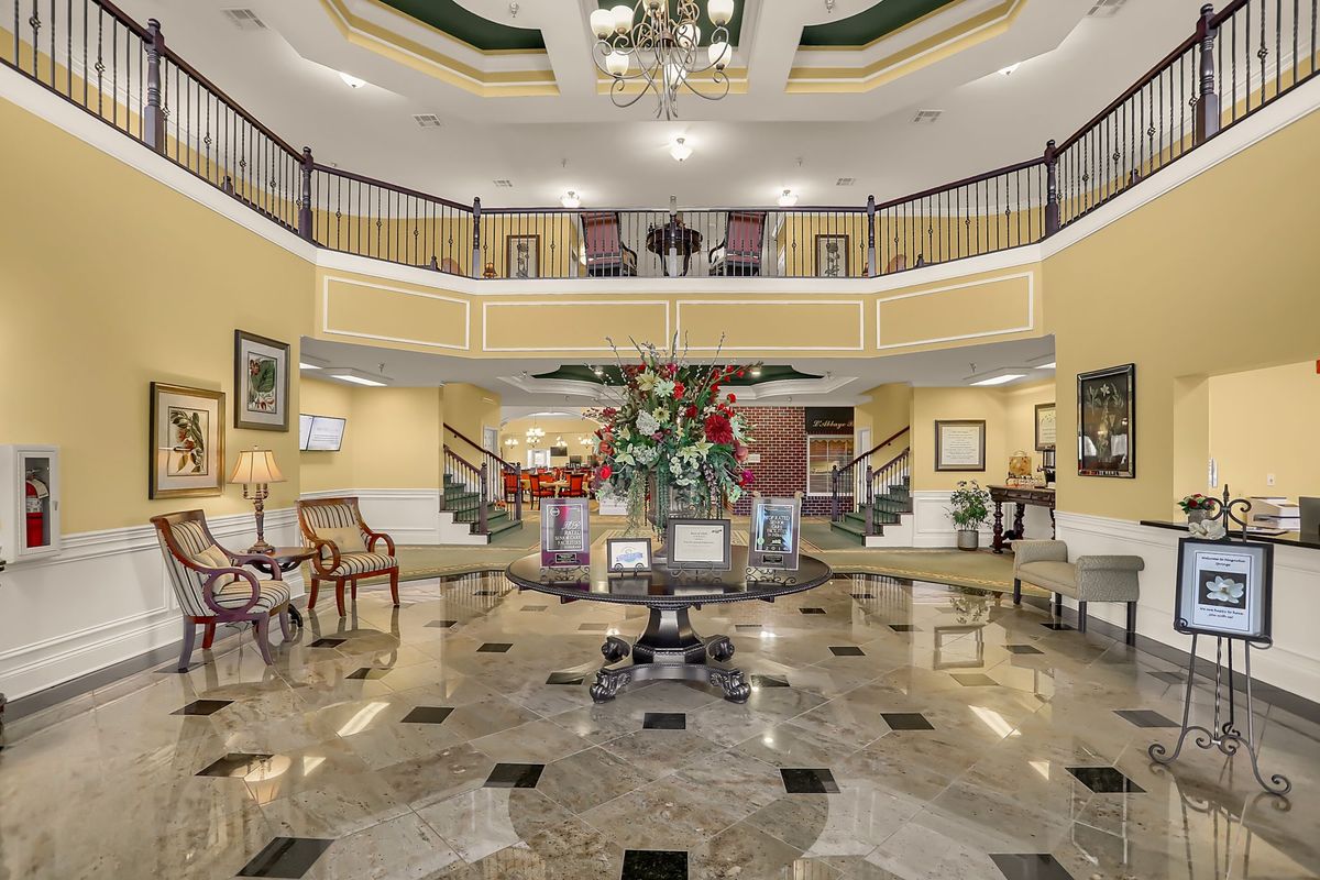 A grand hotel lobby featuring a luxurious interior with a central floral arrangement, elegant staircase, and marble flooring.