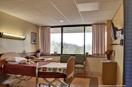 A brightly lit hospital patient room featuring a bed, seating area, and large window overlooking a scenic view.