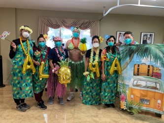 A group of people dressed in Hawaiian-themed costumes pose together with decorations and a banner spelling 'ALOHA'.