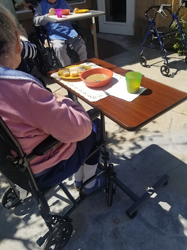 An elderly individual enjoying a meal outdoors, seated in a wheelchair with a tray of food and a drink.