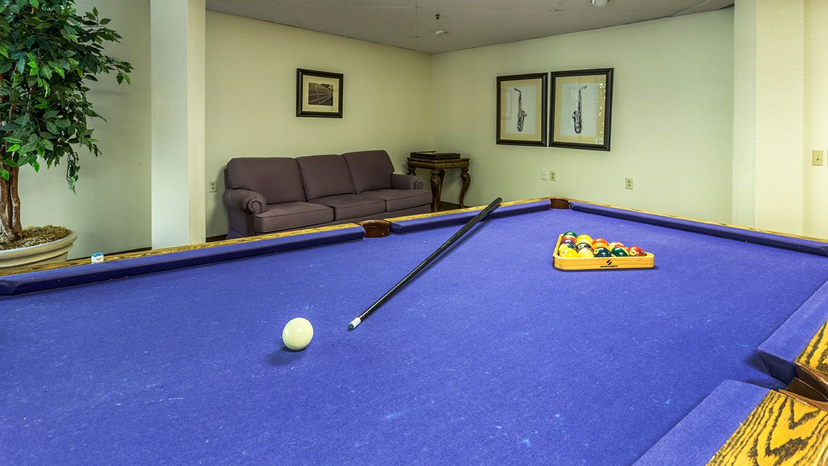 A blue-felt pool table with a cue stick and racked billiard balls in a room featuring a sofa and framed artwork.