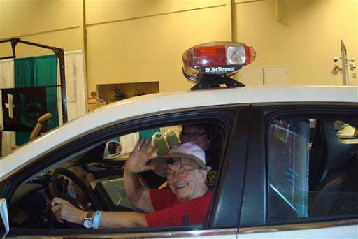 A cheerful individual waves from inside a police car at an event.