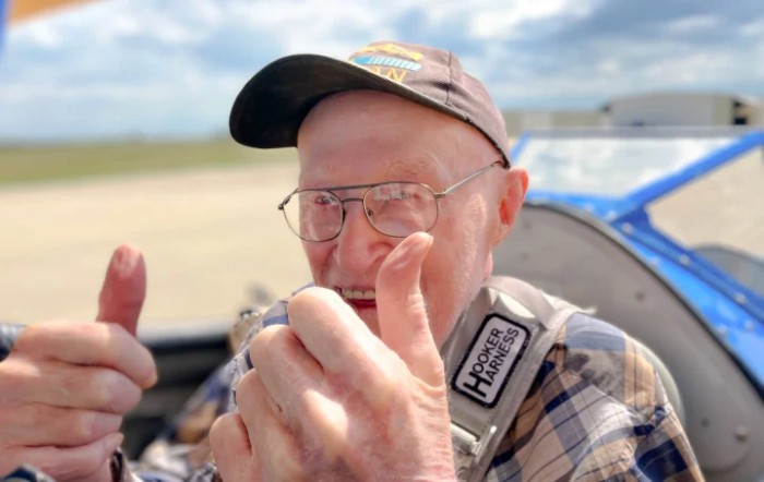 An older man wearing a cap and glasses gives a thumbs up while seated outdoors.