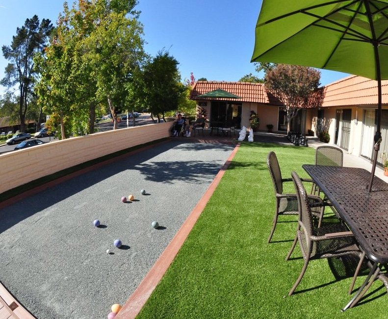 A sunny outdoor area with a bocce court and patio furniture, surrounded by greenery.