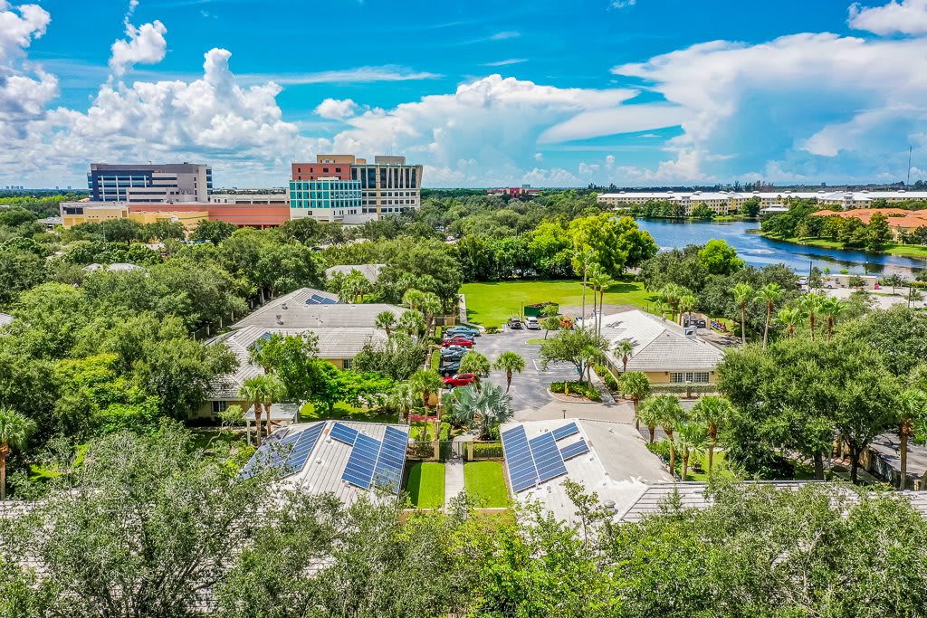 A scenic aerial view highlighting a complex with buildings surrounded by lush greenery and equipped with solar panels, set against a backdrop of clouds and distant structures.