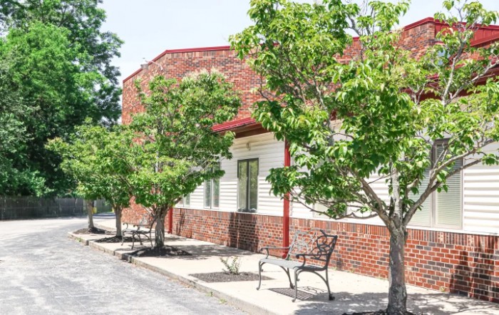 A building with a brick facade is lined with trees and benches along the sidewalk.
