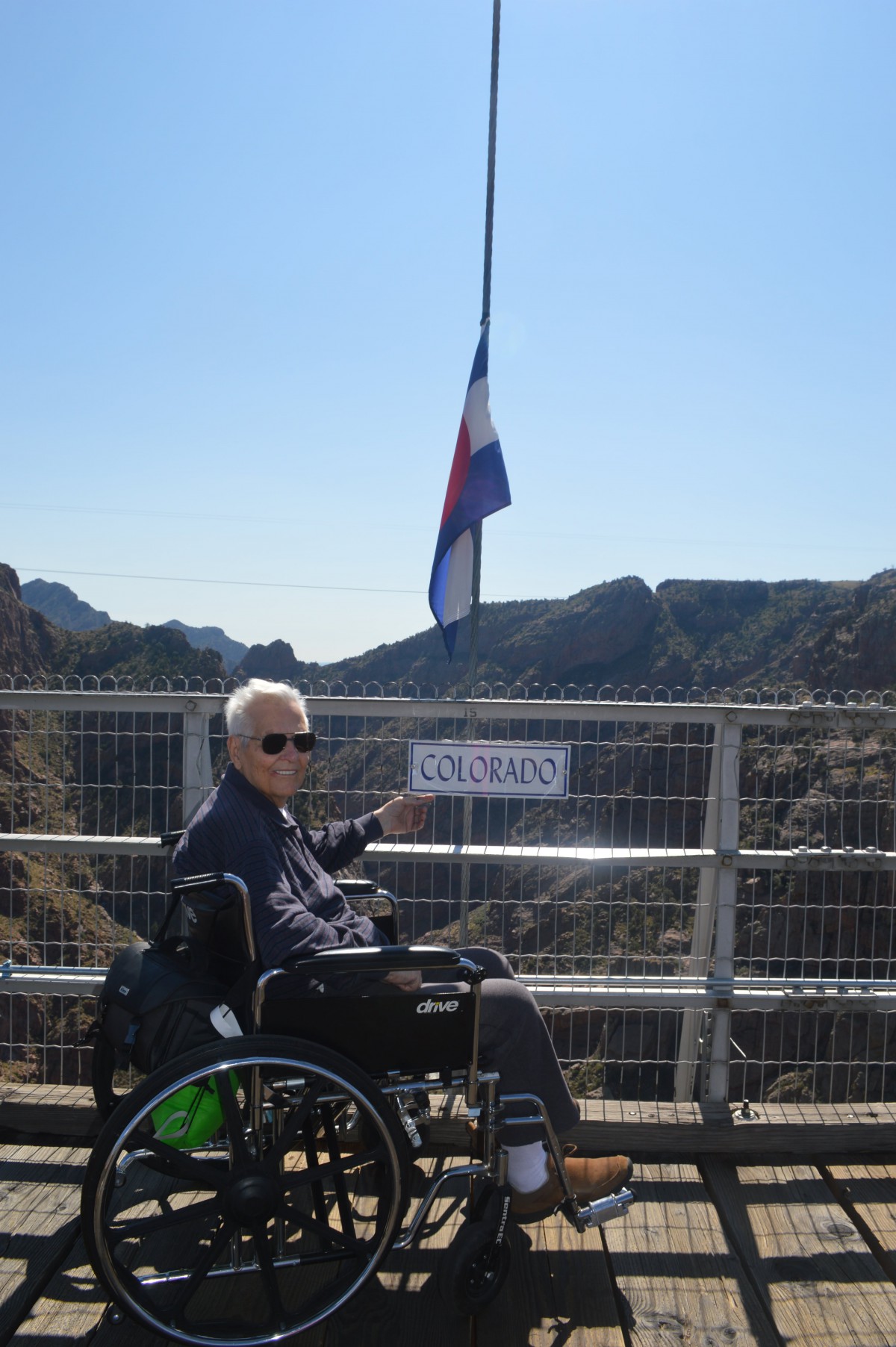 A person in a wheelchair on a bridge with mountains in the background and a flag nearby.