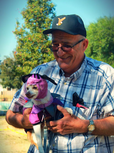 A man happily holds a colorful dog dressed in a costume, enjoying a sunny day outdoors.
