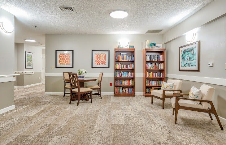 A cozy reading area with bookshelves, chairs, and a small table, featuring framed artwork on the walls.