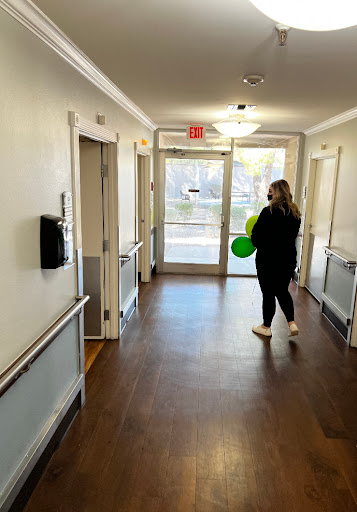A woman holding a green balloon stands in a well-lit hallway with doors on either side.