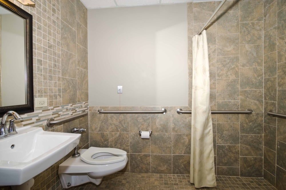 A modern bathroom with tiled walls, featuring a toilet, sink, and shower area with curtain and handrails.