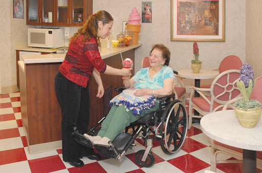A caregiver assists an elderly woman in a wheelchair, bringing her a treat in a cozy indoor setting.