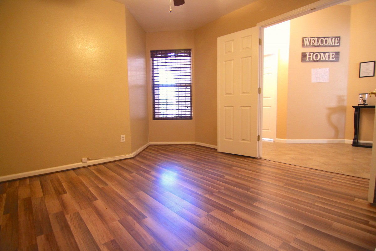 A warm-toned empty room with hardwood flooring, featuring window blinds and an open doorway.