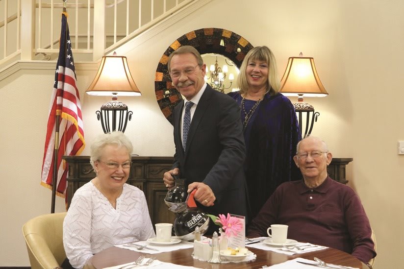 A group of people smiling while one person pours coffee at a table in an indoor setting with a visible American flag.