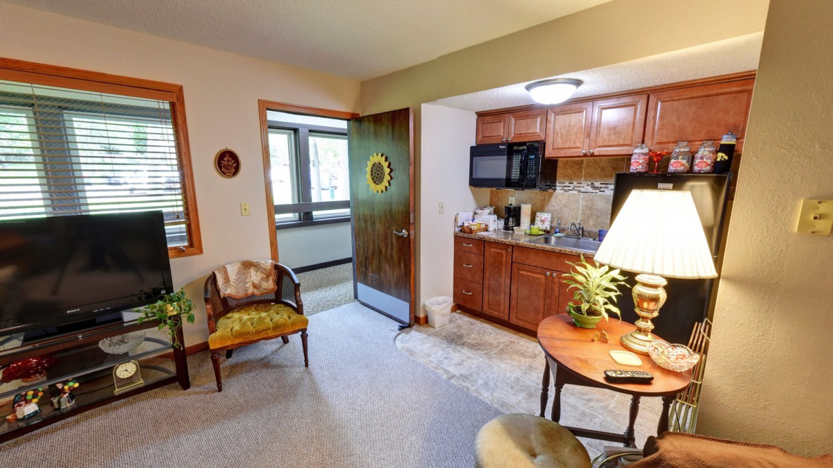 A cozy apartment interior featuring a small kitchen and living area with wooden cabinets, a table with a lamp, and comfortable seating.