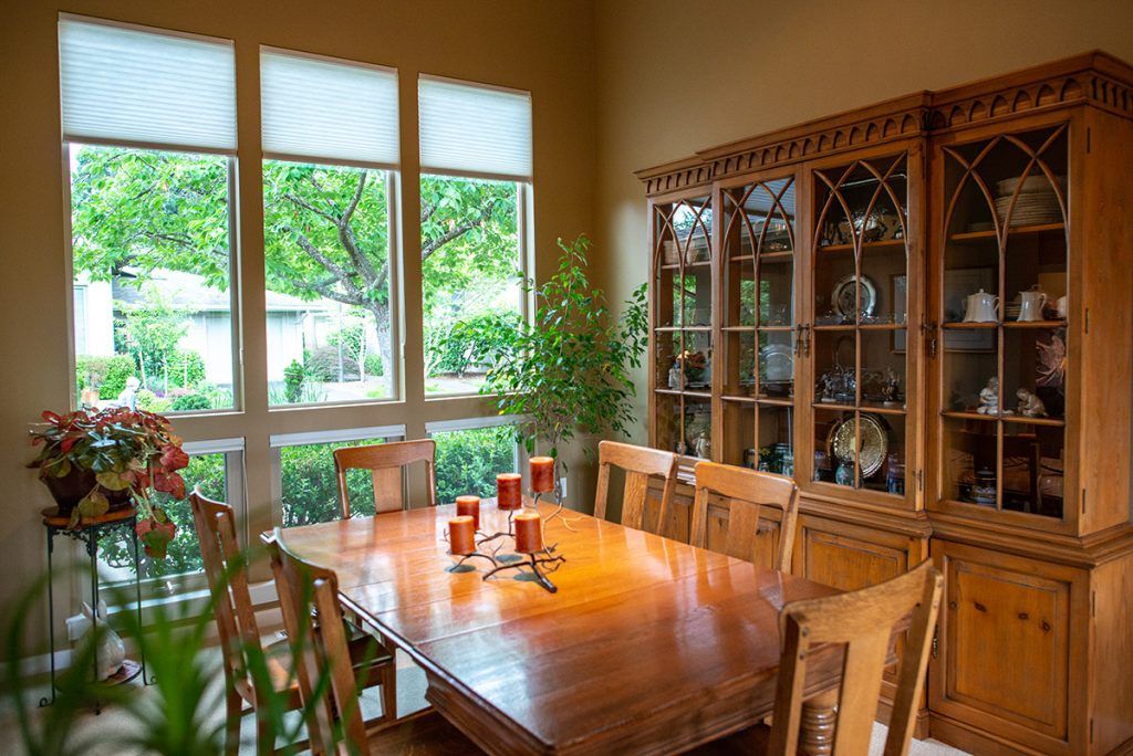 A cozy dining room with a wooden table, surrounded by chairs and featuring a china cabinet. Large windows allow natural light to brighten the space, complemented by indoor plants and candle decorations.