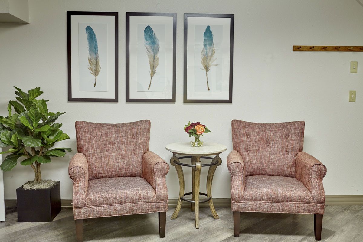 A cozy waiting area with two upholstered chairs, a round table adorned with a small bouquet, and framed feather artwork on the wall.