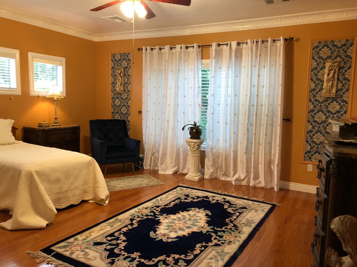 A cozy bedroom with warm tones, featuring a bed, armchair, decorative rug, sheer curtains, and a potted plant.
