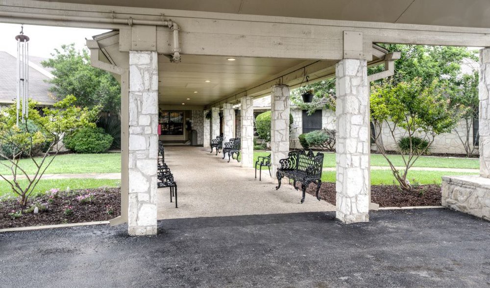 A covered walkway with stone pillars and benches along the sides, surrounded by greenery.