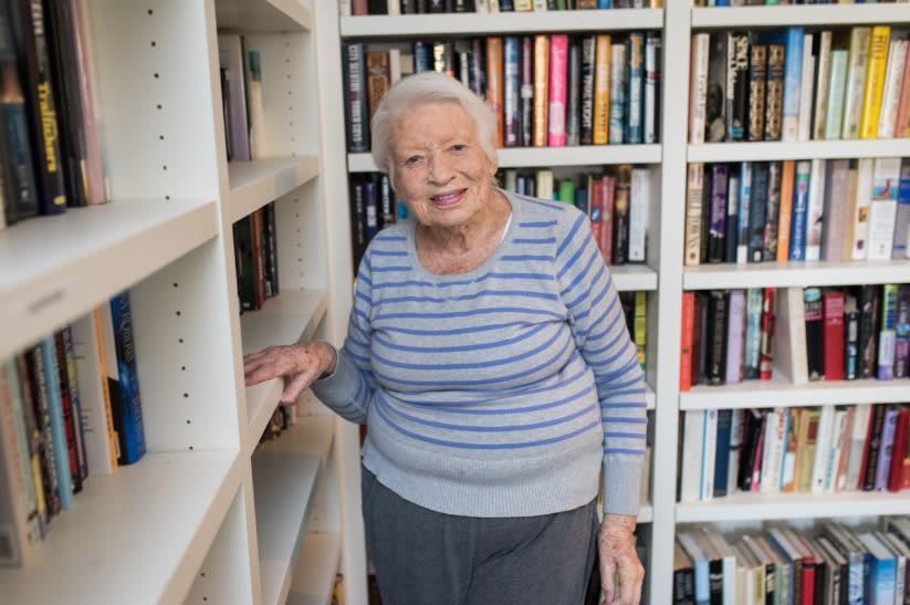 An elderly woman smiling while standing in front of a bookshelf.