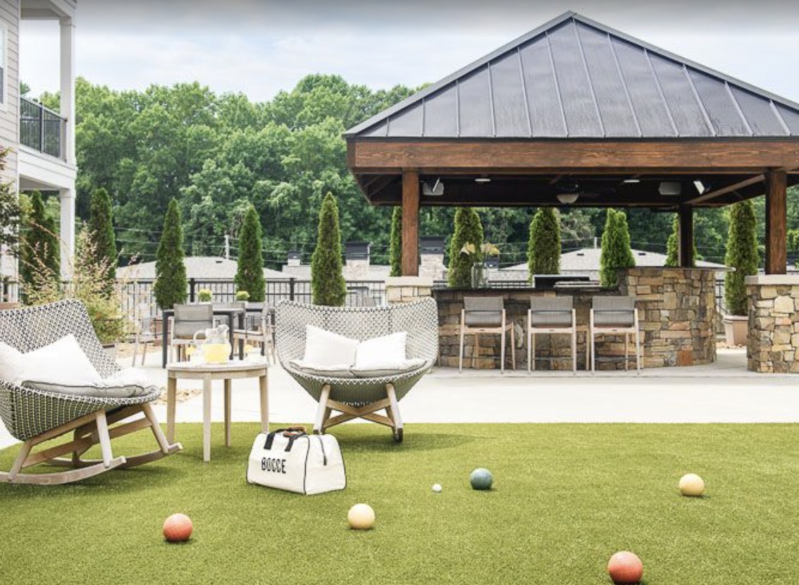 A serene outdoor patio area featuring lounge chairs, a bar space, and bocce balls on a grassy surface, surrounded by lush greenery.