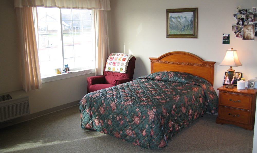 A cozy bedroom featuring a single bed with floral bedding, an armchair by the window, and a landscape painting on the wall.