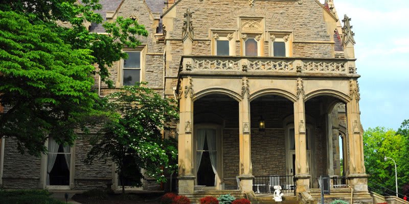 A historic stone mansion with arched windows and a porch featuring columns, surrounded by a garden.