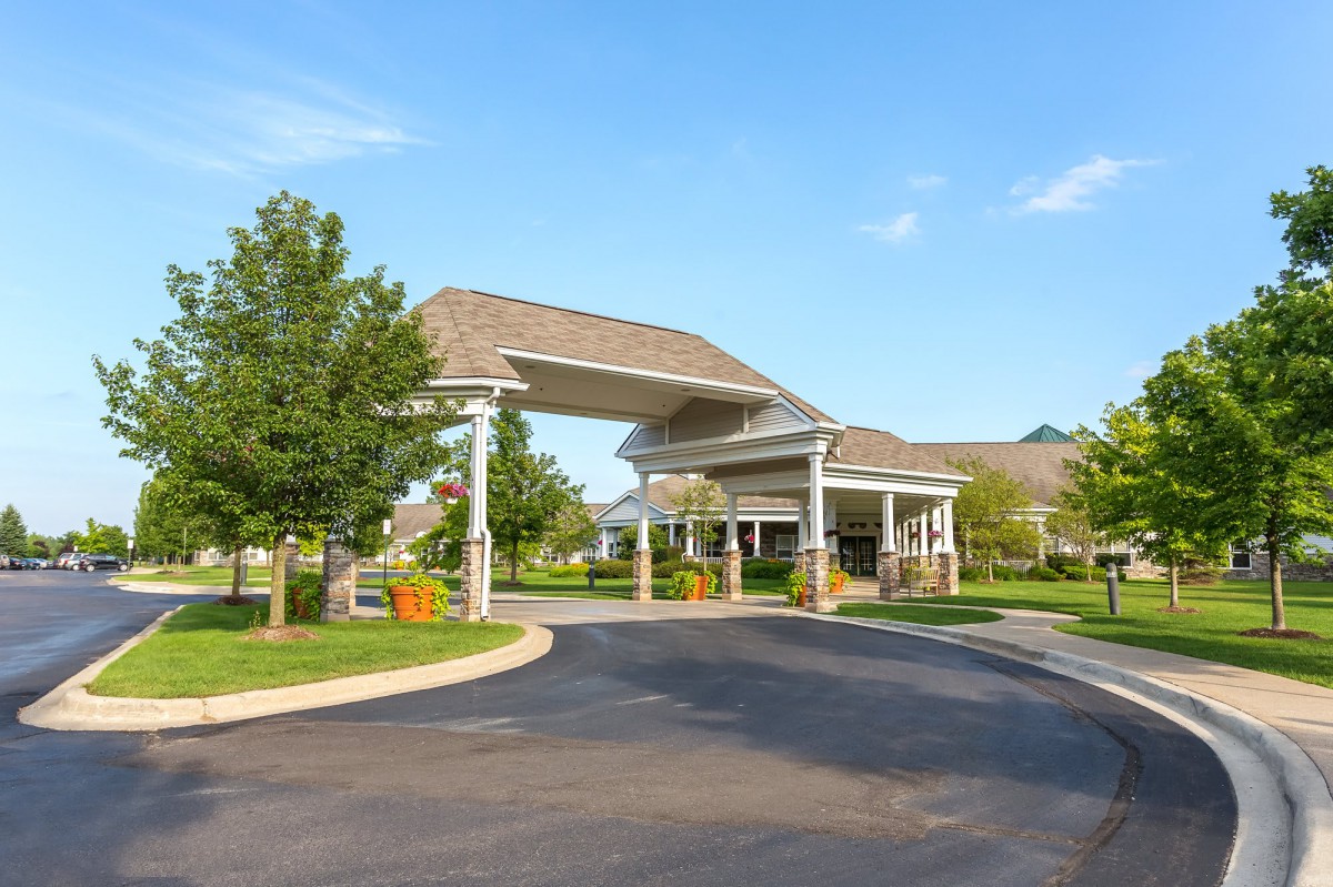 A sunny view of a building entrance with a landscaped driveway and surrounding greenery.