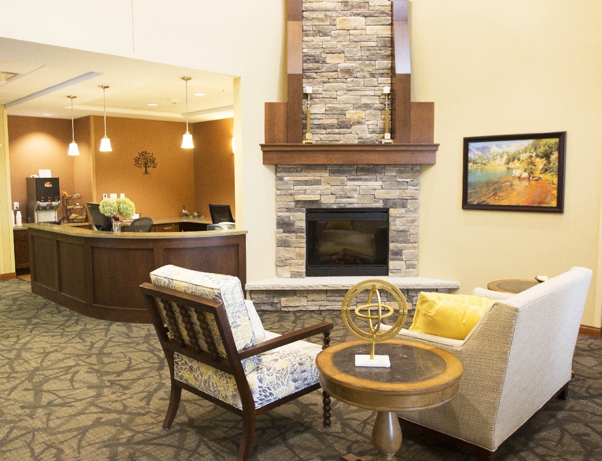 The image shows a cozy lobby area with a stone fireplace, comfortable seating, and a reception desk in the background.