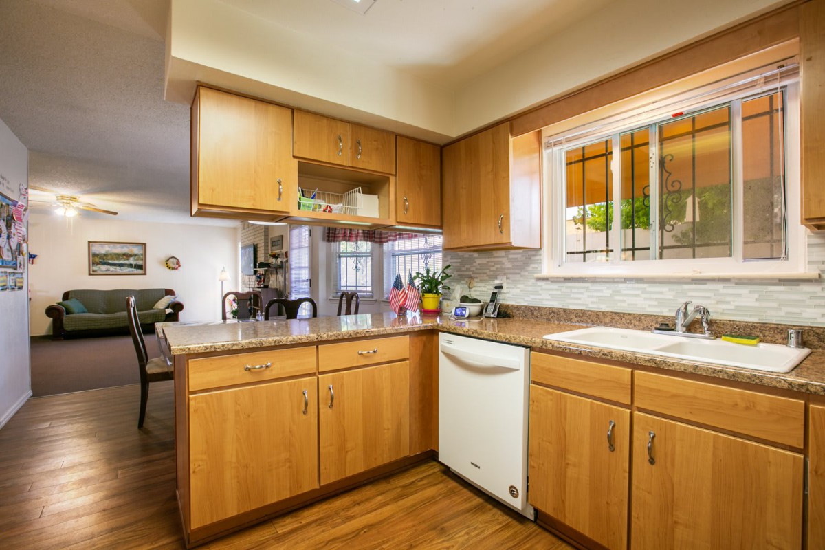 A modern kitchen with wooden cabinets and a dishwasher under a countertop, featuring a window and hardwood flooring.