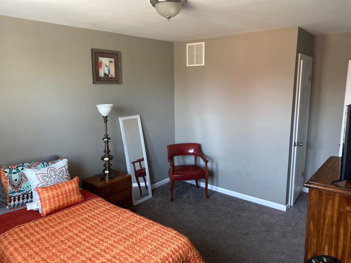 A minimalist bedroom with orange bedding, a standing lamp, a decorative mirror, and a red chair.
