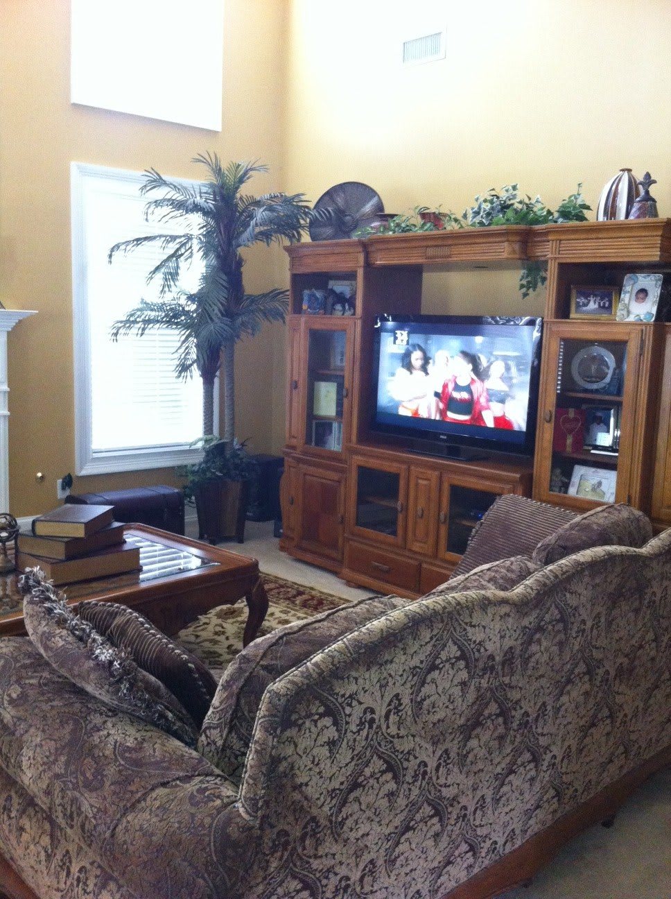 A cozy living room with a large entertainment center and television, featuring a patterned couch and a coffee table set against warm yellow walls.