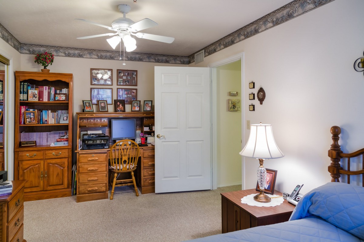 A cozy bedroom with a wooden desk, chair, and computer setup, illuminated by a table lamp. A ceiling fan hangs from the ceiling.
