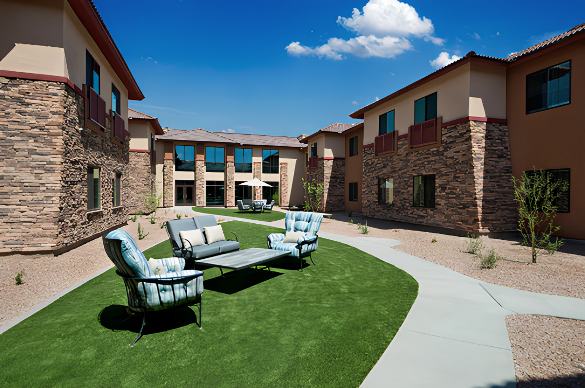 A sunny courtyard with outdoor seating, featuring a sofa and chairs on a grassy area surrounded by buildings.