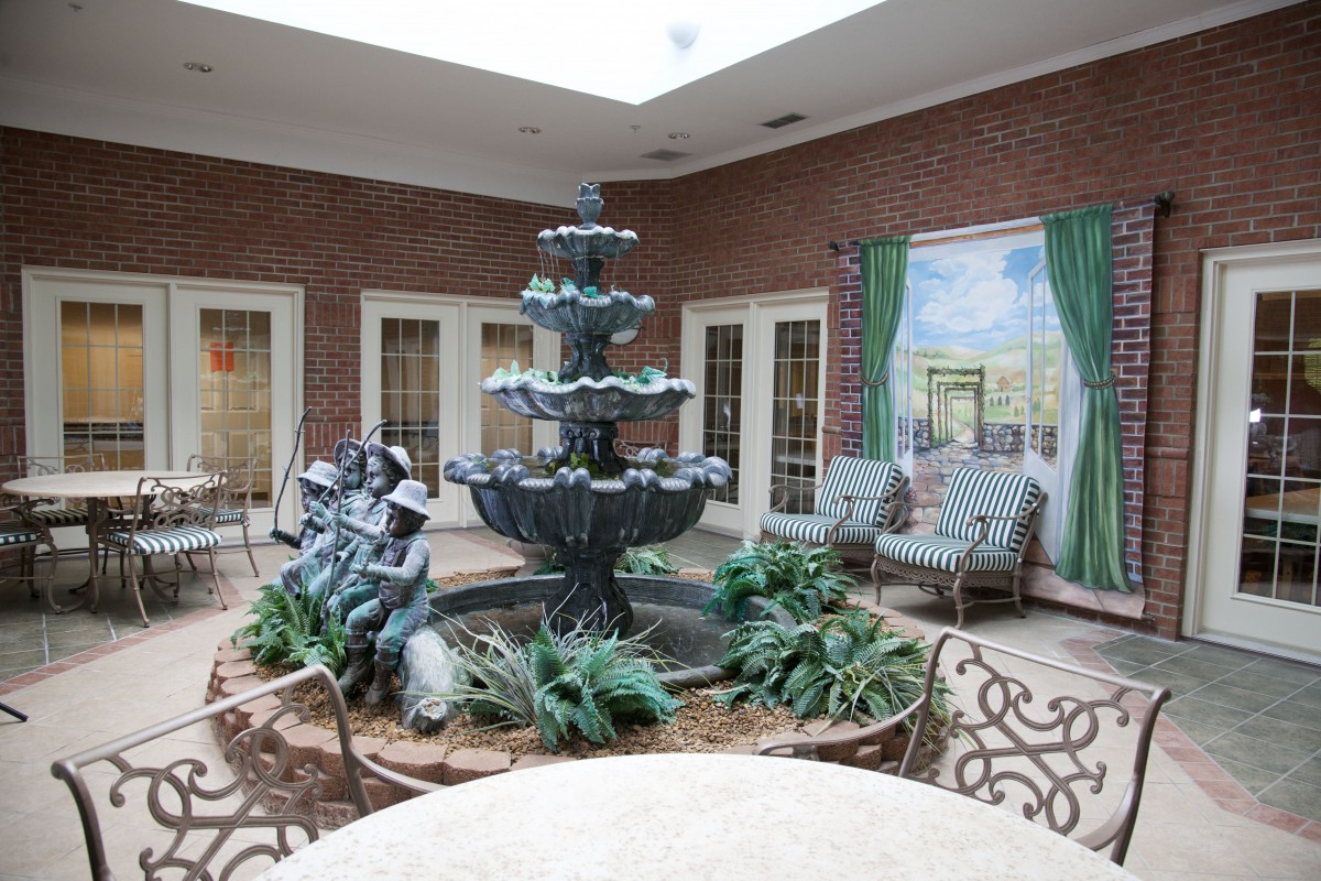A serene indoor courtyard featuring a multi-tiered fountain surrounded by statues, plants, and comfortable seating in a well-lit space with brick walls.