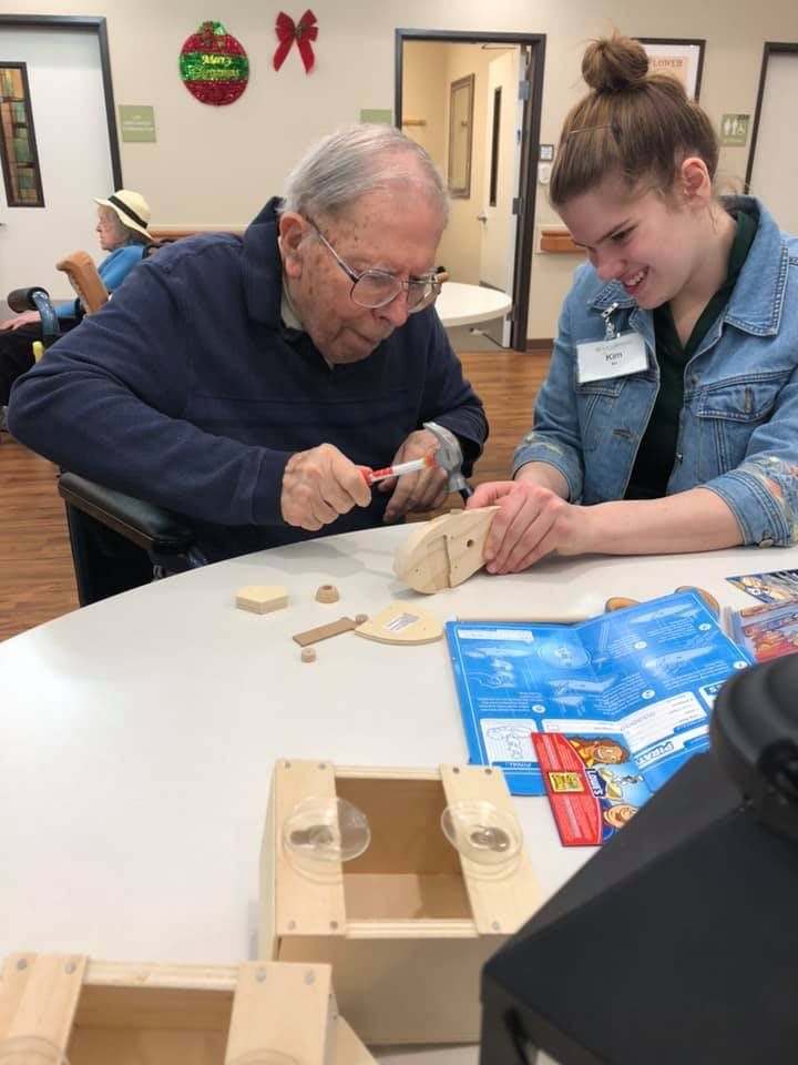 An elderly man works on a woodworking project with assistance from a young woman.