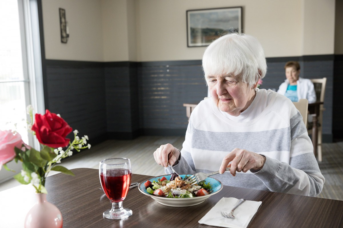 An elderly woman enjoys a meal at a table set with a salad and a glass of red drink.