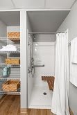 A compact bathroom featuring a shower area with a curtain, adjacent to shelves stocked with towels and baskets.