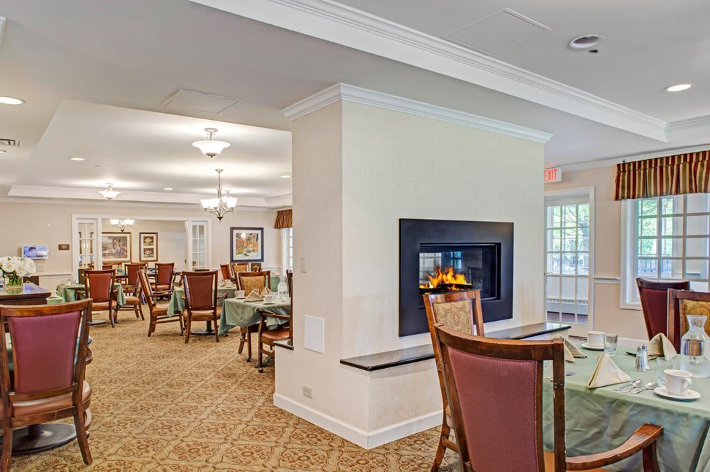 A cozy dining area with a central fireplace, surrounded by tables and chairs, featuring warm lighting and carpeted flooring.