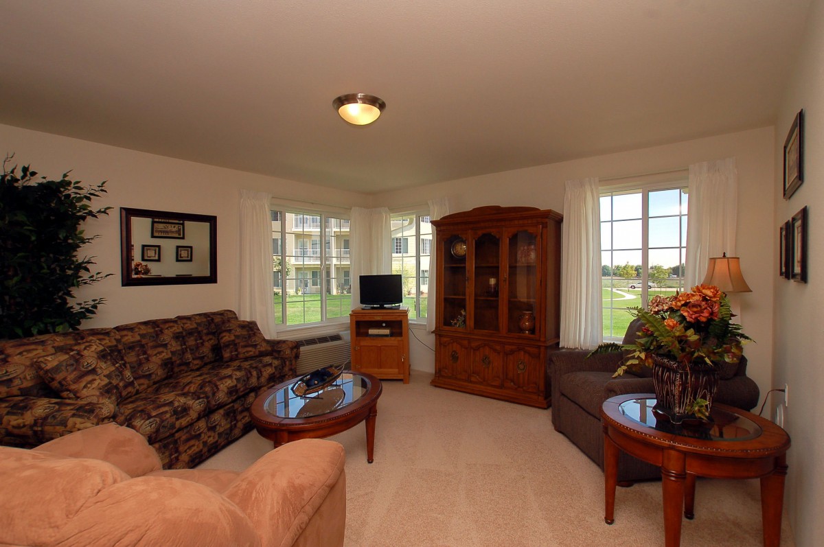 A cozy living room featuring sofas, armchairs, a television, and a wooden cabinet in a well-lit space with large windows.