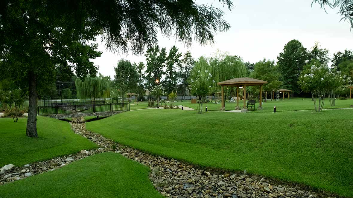 A serene park landscape featuring a gazebo, lush grass, a small creek, and surrounding trees.
