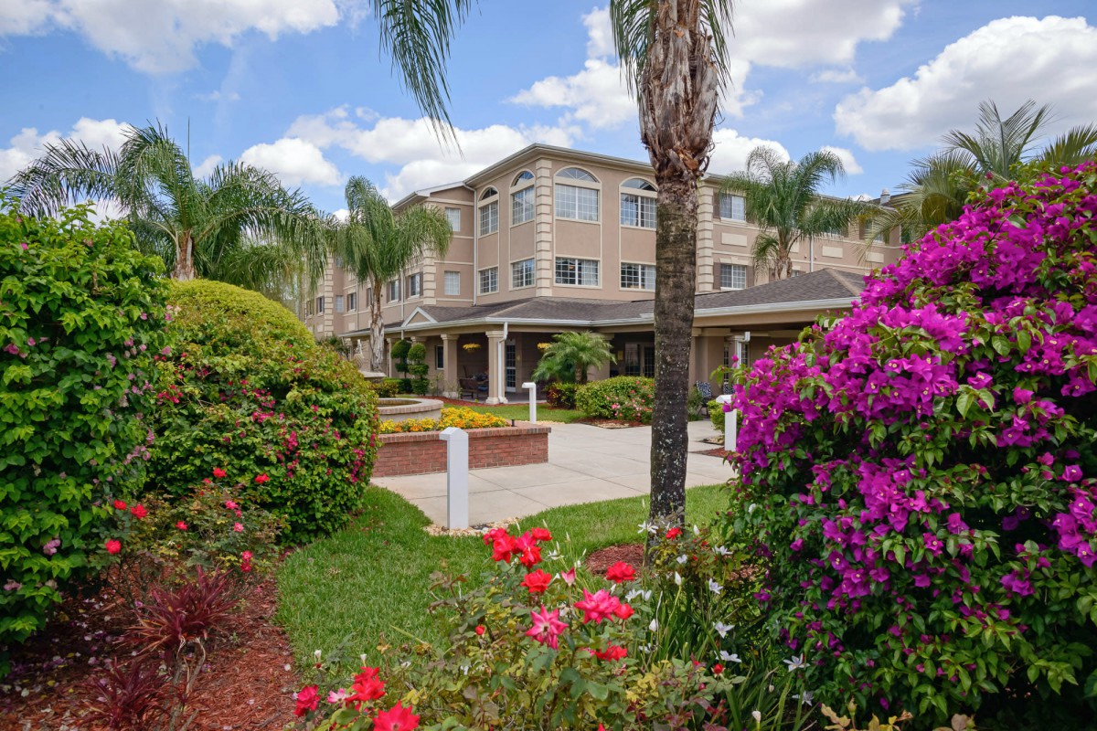 A residential building surrounded by a beautifully landscaped garden with vibrant flowers and palm trees under a partly cloudy sky.