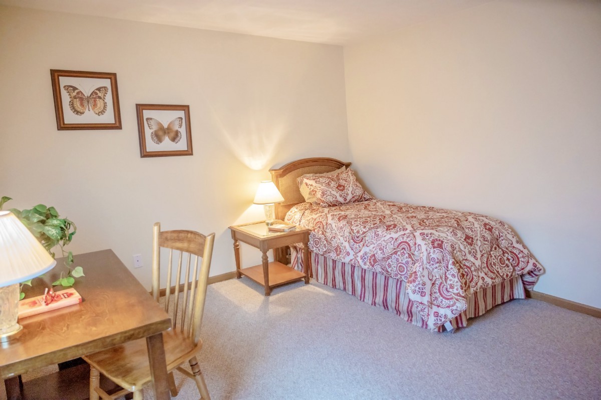 A cozy bedroom with a single bed, side table, lamp, and a wooden desk with a chair.